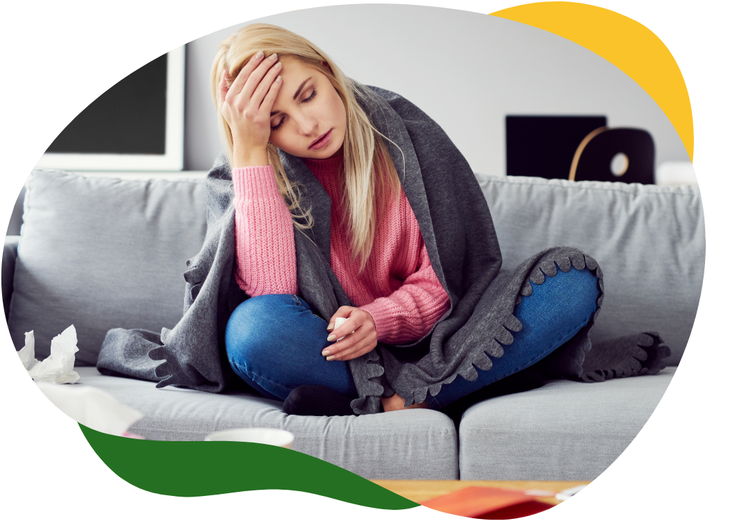 A young woman sits cross-legged on the sofa with her back bent and her right arm supporting her forehead. She is covered with a blanket