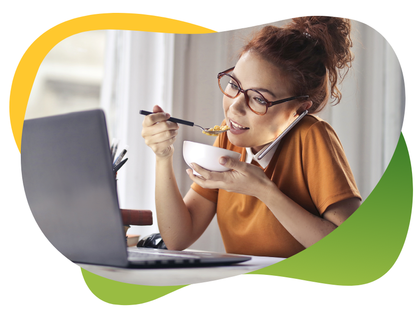 Young woman with glasses and high braid sits in front of laptop while talking on phone and eating her meal at the same time