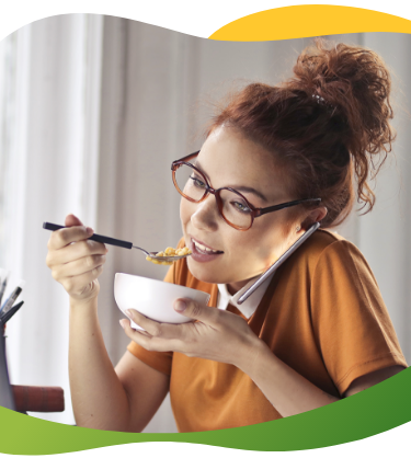 Young woman with glasses and high braid sits in front of laptop while talking on phone and eating her meal at the same time